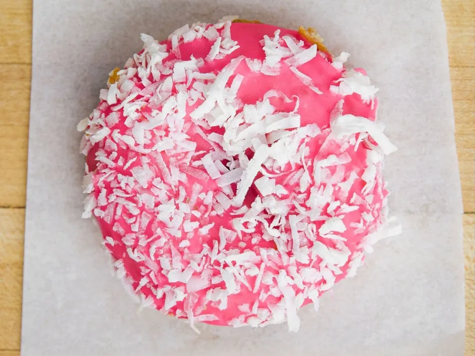 A donut with pink glaze and shredded coconut, on white paper.