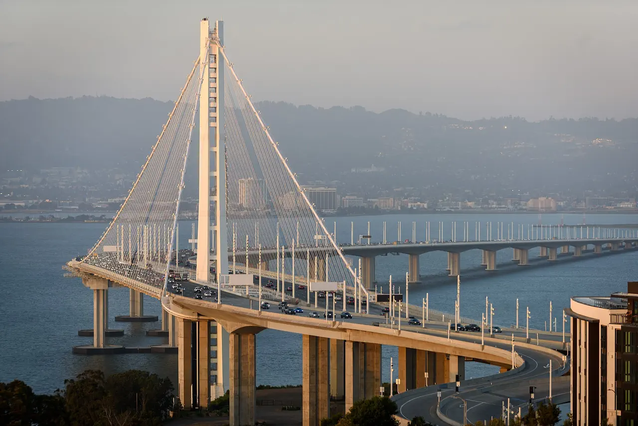 The Bay Bridge's white tower and curving eastern span, busy with traffic, seen from Yerba Buena Island with the Oakland shoreline and hills behind.