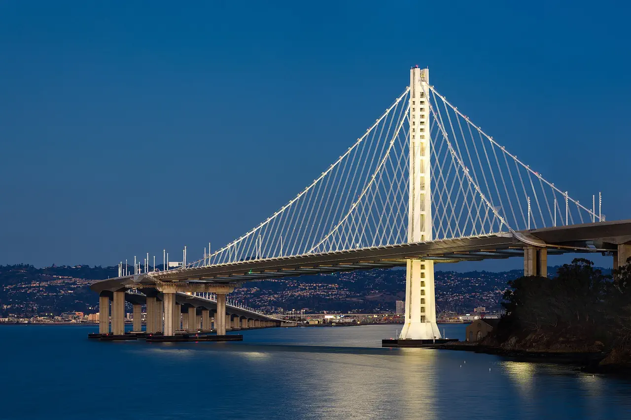 The lit tower and cables of the Bay Bridge's eastern span against a deep blue evening sky, with shoreline lights across the water.