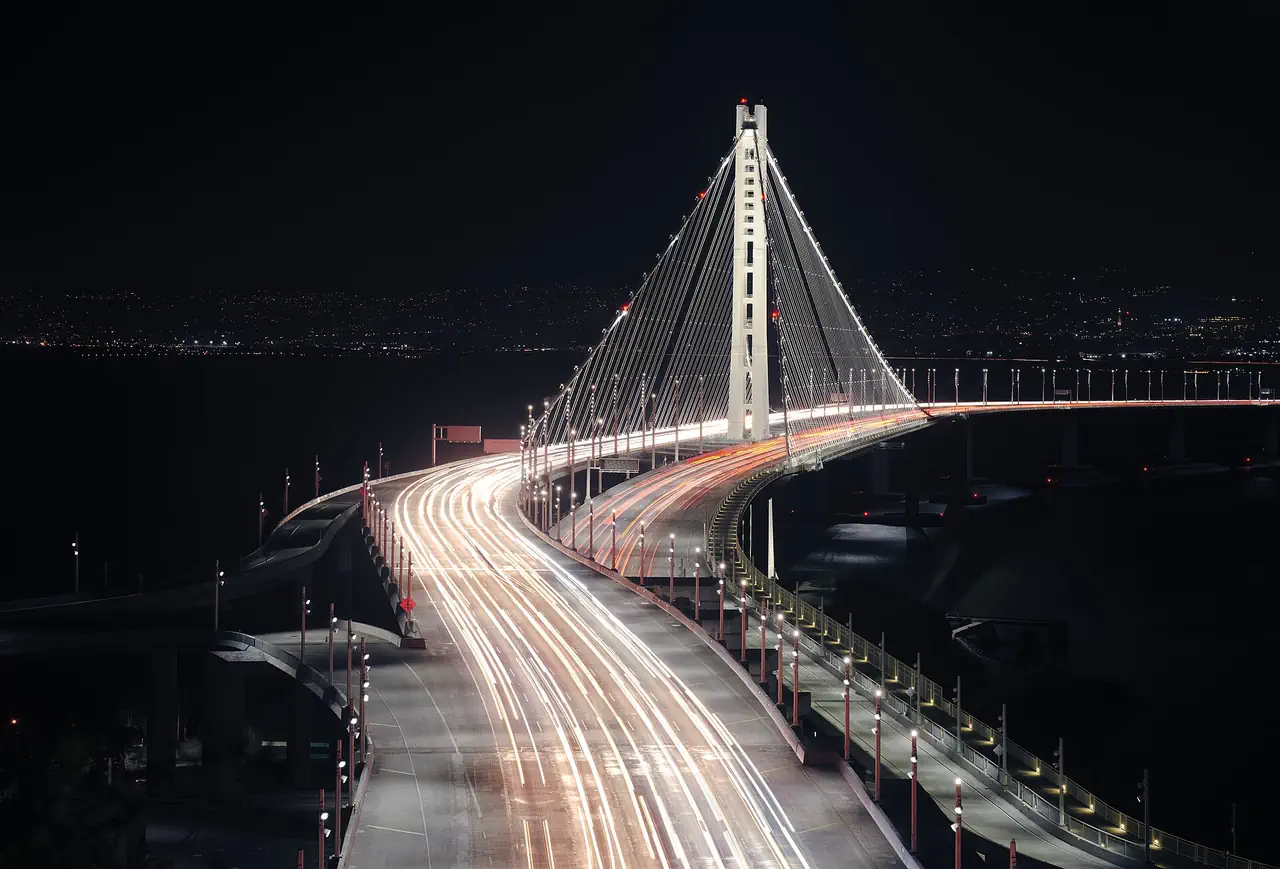 Light trails from traffic curving across the Bay Bridge's eastern span at night, seen from Yerba Buena Island, with city lights across the bay.