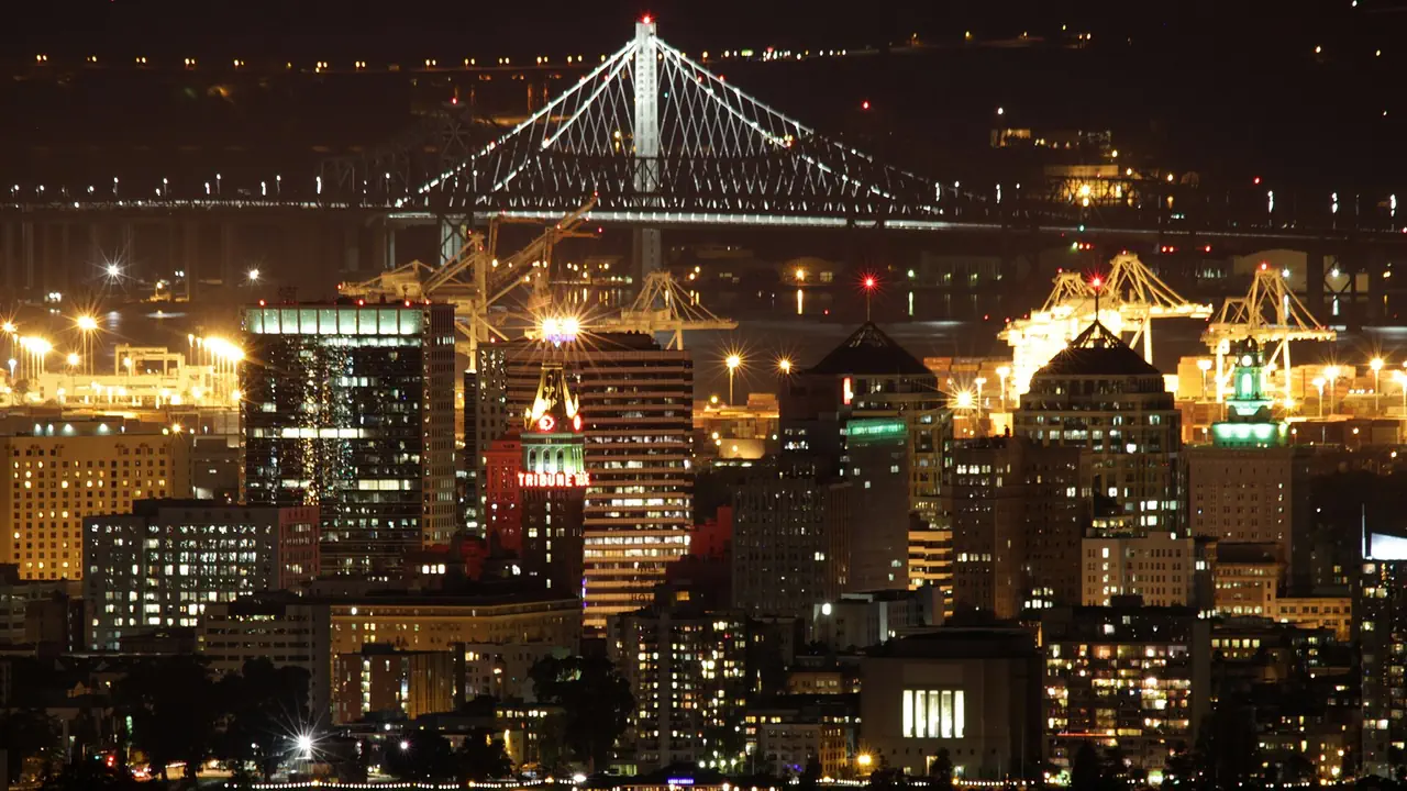 Downtown Oakland lit up at night, with the Tribune Tower, the port's cranes and the Bay Bridge's eastern span glowing behind the skyline.