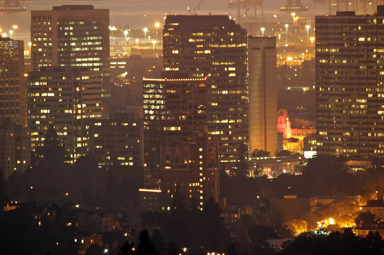 Office towers in downtown Oakland with their windows lit at dusk, and port cranes in the haze beyond.