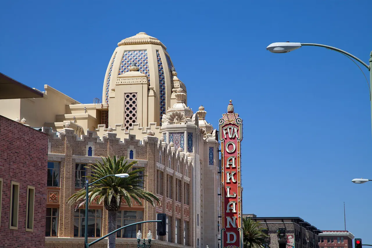 The Fox Oakland Theatre's tiled dome and tall red FOX OAKLAND sign rising above palm trees and street lamps in Uptown Oakland, under a clear blue sky.