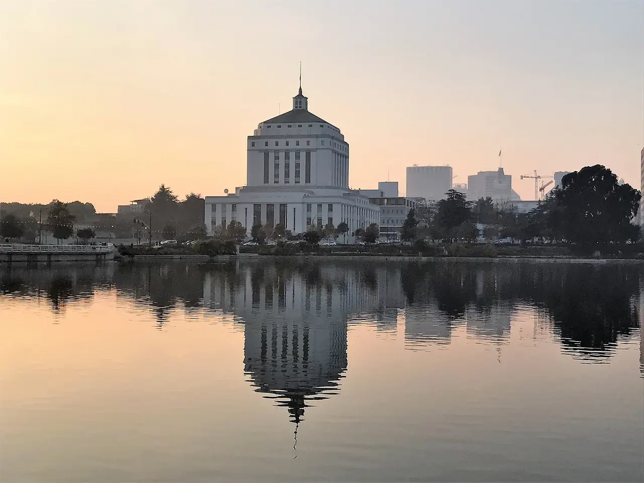 The Alameda County Courthouse reflected in Lake Merritt, Oakland, under a hazy peach sky near sunset.