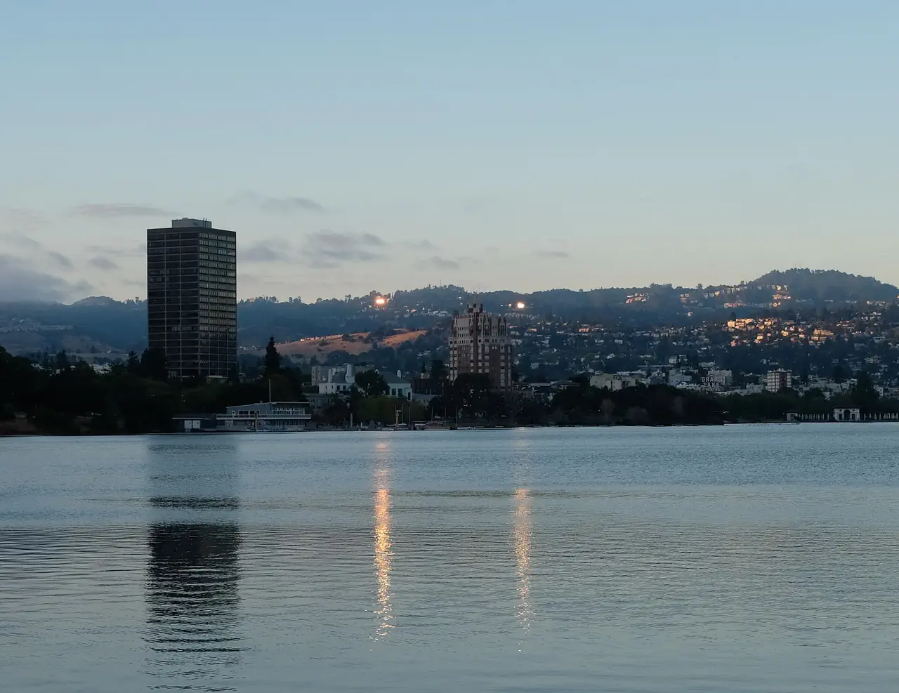 Lake Merritt, Oakland, at dusk, with high-rises along the shore and the Oakland hills beyond the still water.