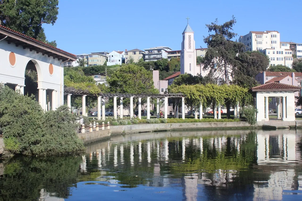 The white colonnade and pergola on the Grand Avenue side of Lake Merritt, Oakland, reflected in the water, with a church tower and hillside apartments behind.