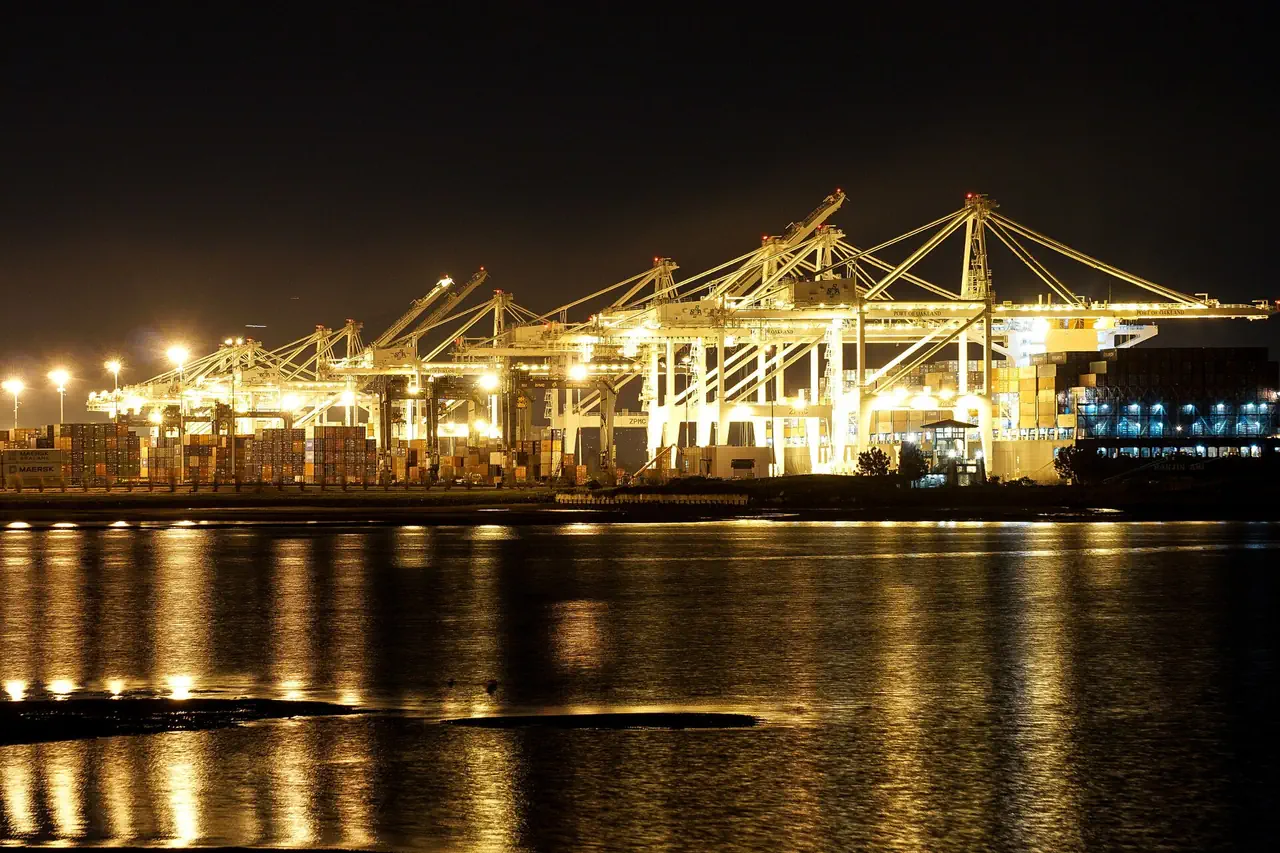 Brightly lit container cranes and stacked containers at the Port of Oakland at night, reflected in the dark water.
