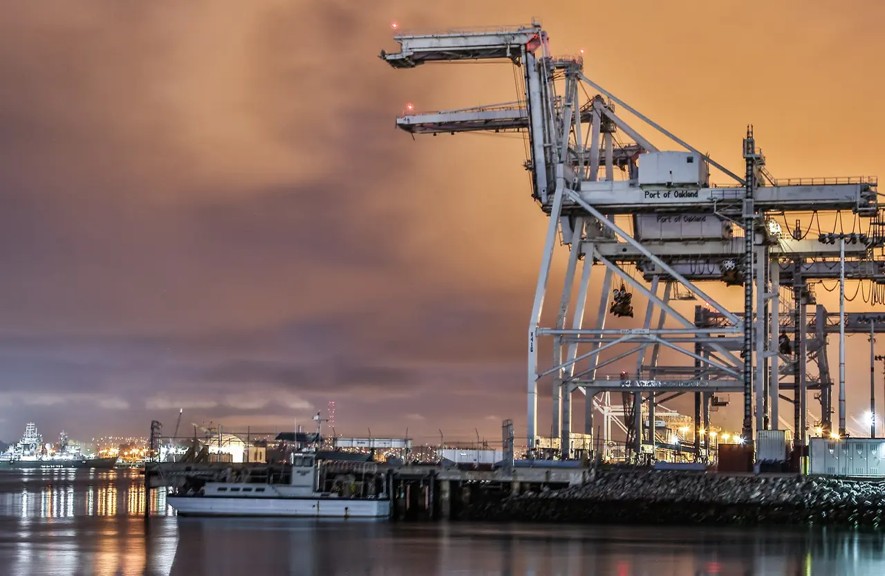 Port of Oakland container cranes under low clouds glowing orange in the dark, with a small boat moored at the pier below.