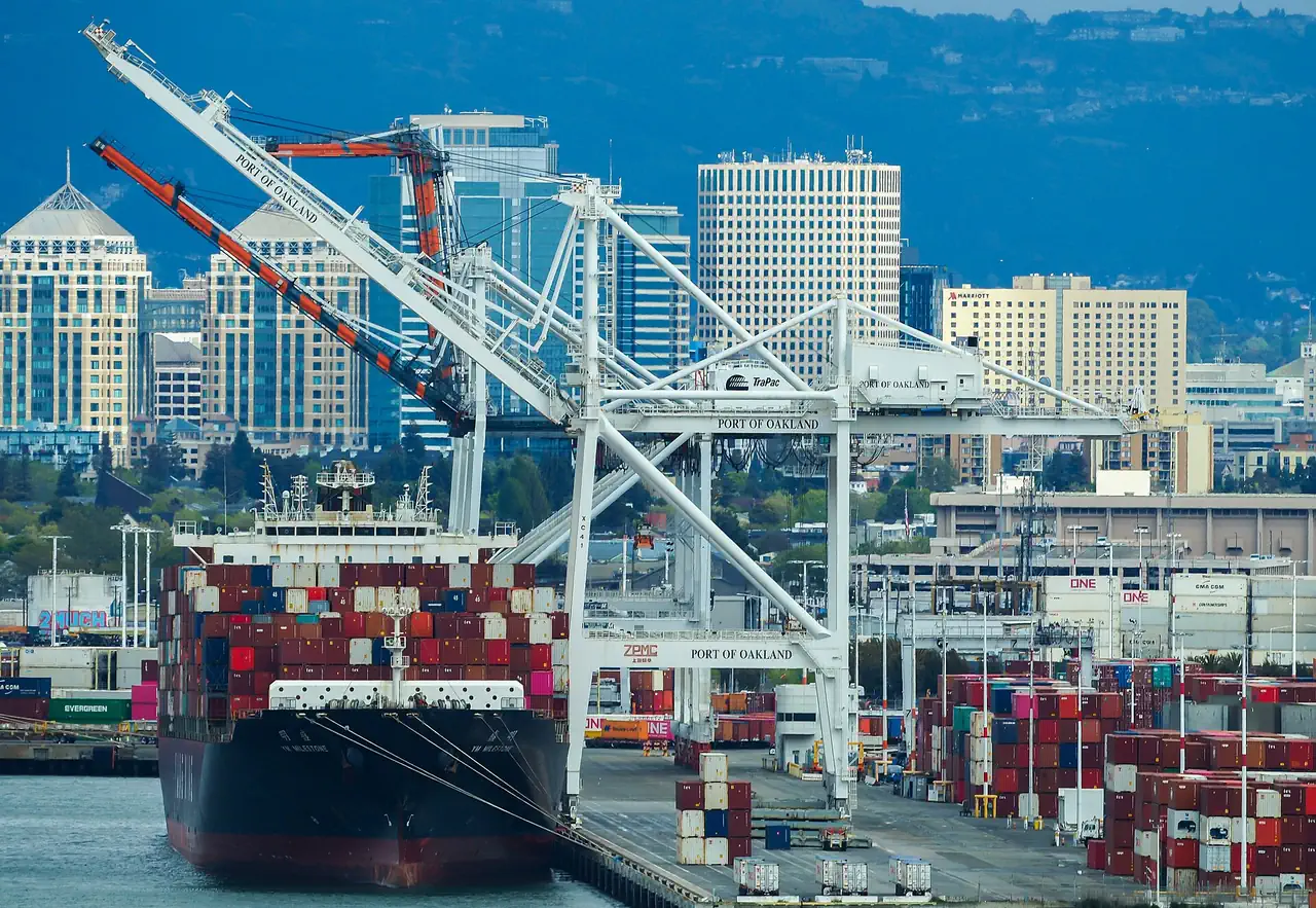 A loaded container ship docked beneath Port of Oakland cranes, with downtown Oakland's towers and the hills behind.