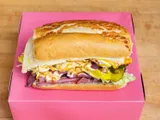 A pastrami sandwich on a roll with cheese, lettuce and peppers, on a pink box.