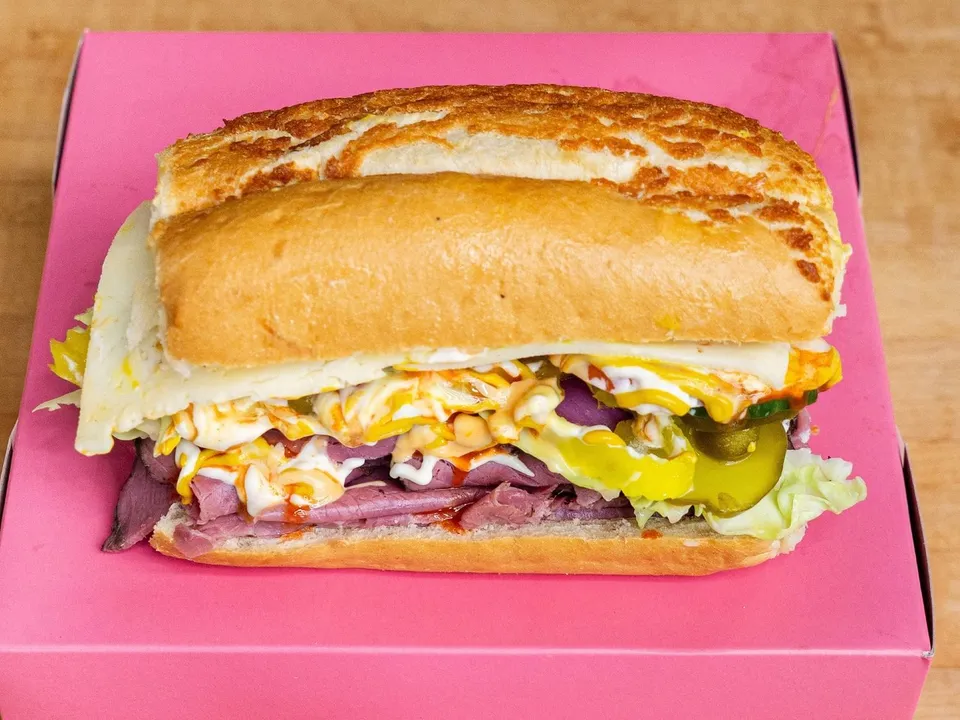 A pastrami sandwich on a roll with cheese, lettuce and peppers, on a pink box.