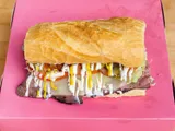 A roast beef sandwich on a roll with cheese, tomato, lettuce and pickles, on a pink box.