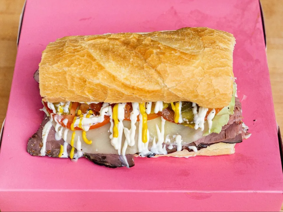 A roast beef sandwich on a roll with cheese, tomato, lettuce and pickles, on a pink box.