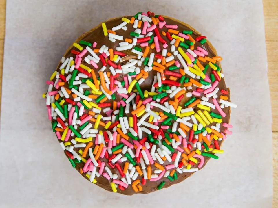 A cake donut with chocolate glaze and rainbow sprinkles, on white paper.